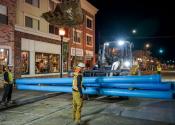 A construction worker guides heavy equipment carrying blue pipe during a nighttime job.